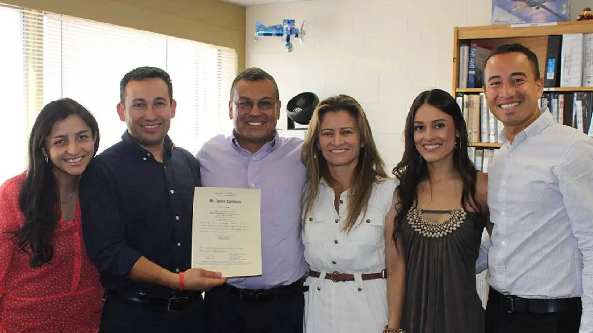 A group of six people posing together, with one individual holding a certificate, in a brightly lit room with bookshelves.