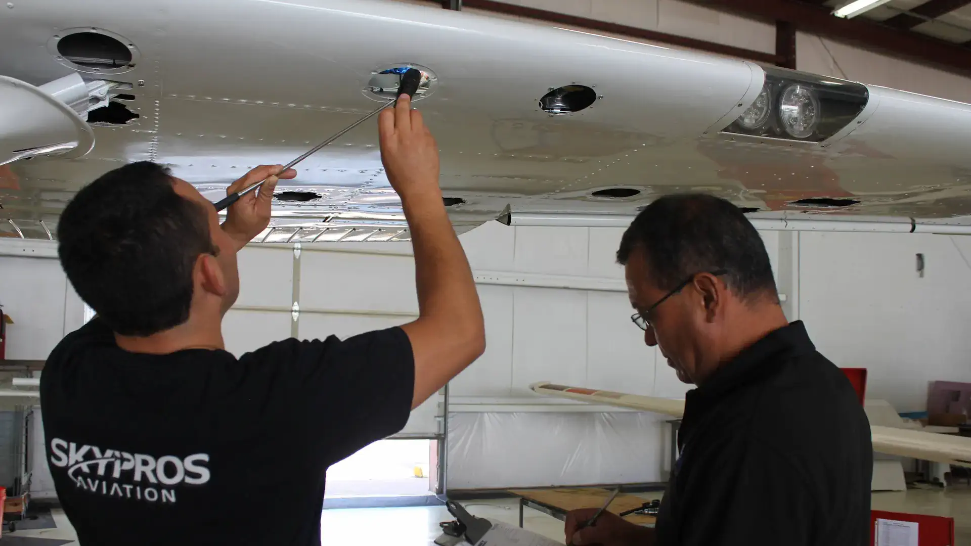 Two technicians inspect an aircraft's wing, using tools to check components in a well-lit hangar.